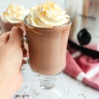 hand holding up a cup of hot chocolate with a second cup in the background with crockpot