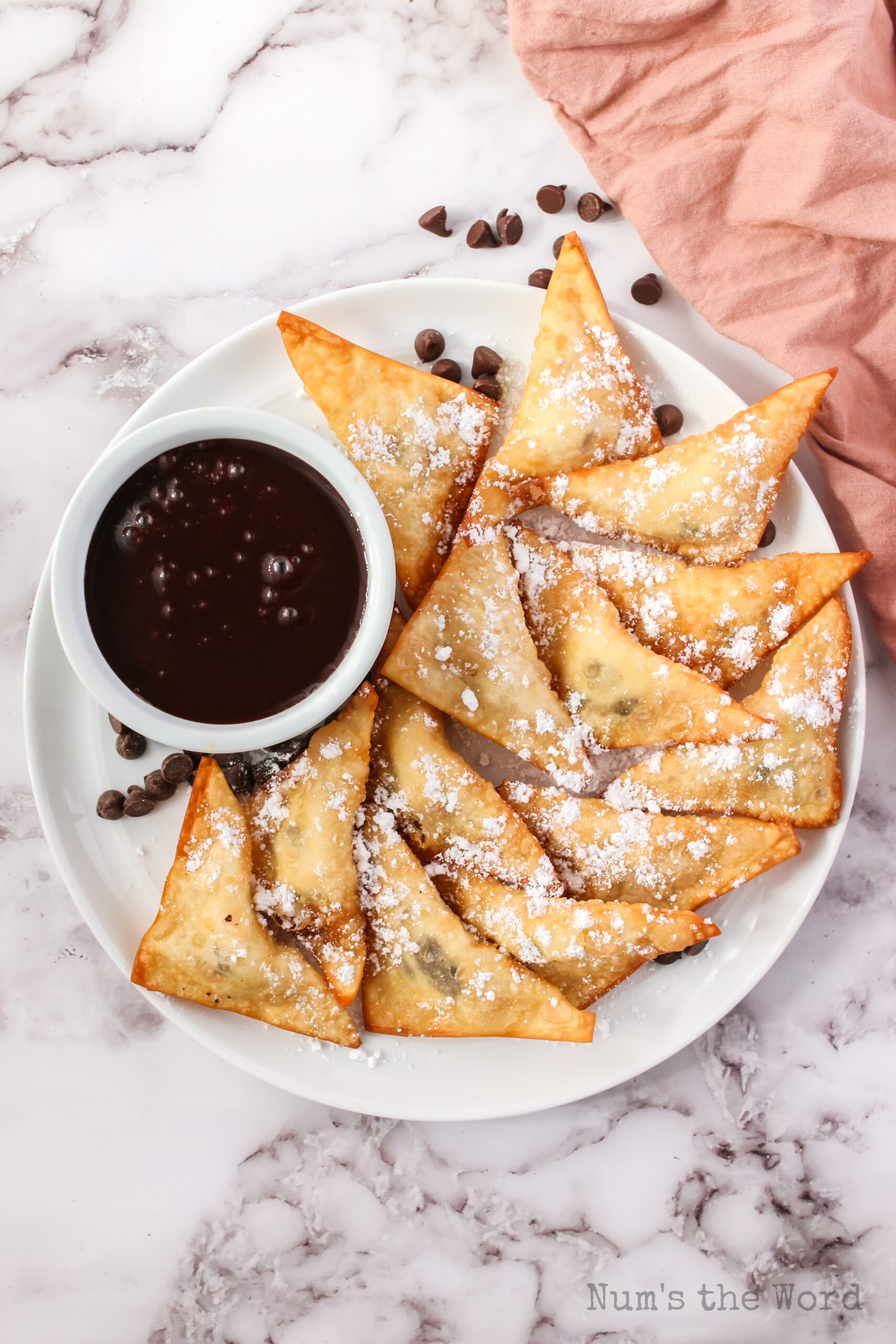 plate full of cooked dessert wontons sprinkled with powdered sugar with a bowl of chocolate sauce
