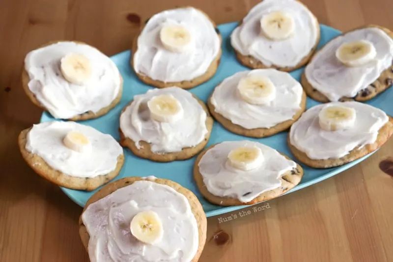 Banana Chocolate Chip Cookies - platter of frosted cookies with frosting an a slice of banana on top. One cookie is in front propped up on plate.
