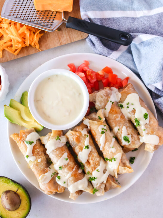 6 chimichangas on plate with sauce drizzled over the top. Avocados and tomatoes on plate as well.