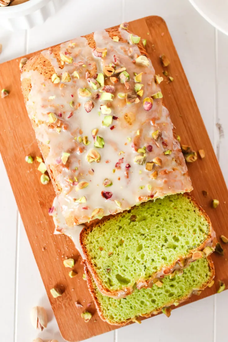 A loaf of bread on a cutting board with almond icing and chopped nuts drizzled on top. Two slices have been cut off.