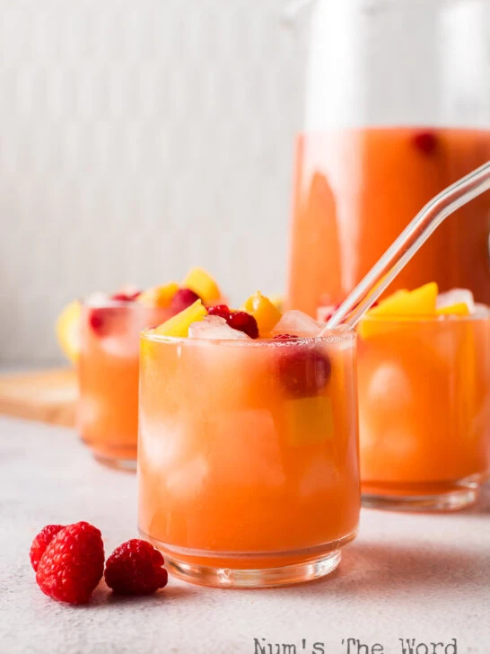 side view of 2 drinks in glasses with a pitcher of lemonade in the background.