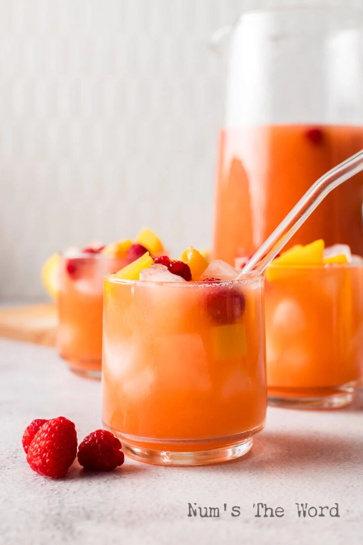 side view of 2 drinks in glasses with a pitcher of lemonade in the background.