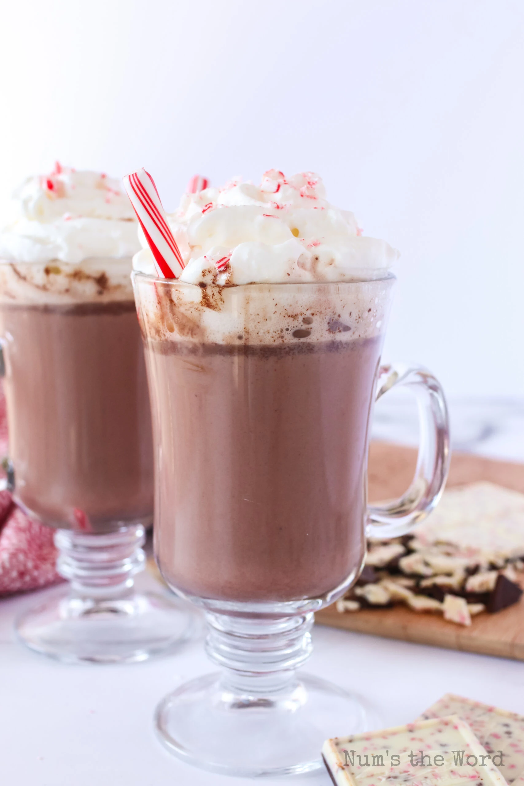 side view of 2 glasses of hot cocoa with whipped cream, a peppermint stick and crushed peppermint on top