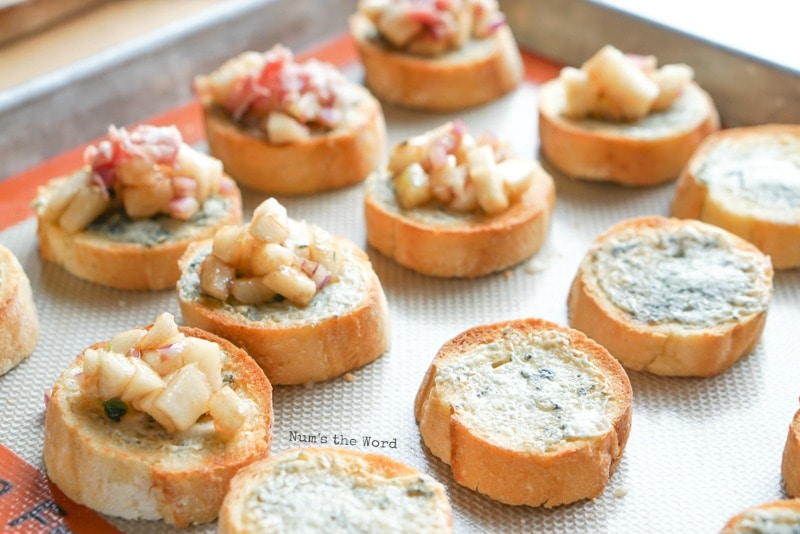 Pear & Proscuitto Bruschetta - french bread bites on cookie sheet, showing the different stages, one row with melted blue cheese on bread bites, another row with melted blue cheese and pear mixture and another row with blue cheese, pear mixture and prosciutto.