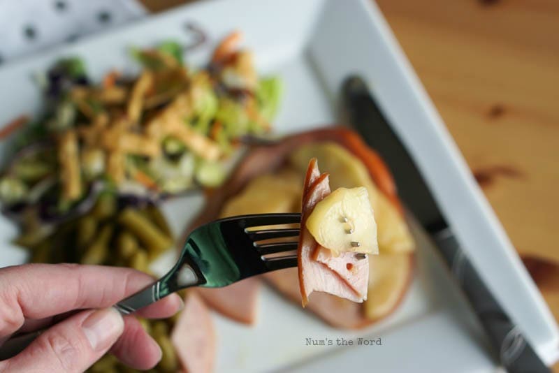 Ham Steaks with Sauteed Apples - A bite of ham and apples on a fork close to camera with plate full of food in background