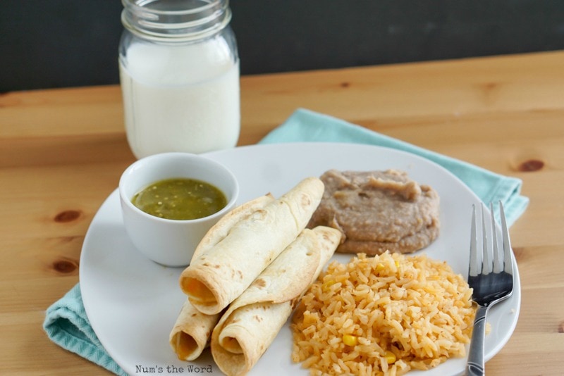 Pulled Pork Flautas - 3 tortillas on a plate with refried beans and mexican rice. Zoomed out photo includes a small dish of salsa verde too.