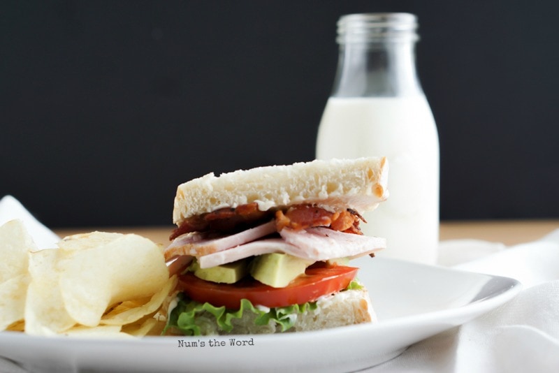 Roasted Turkey & Avocado BLT - Sandwich on a plate with chips. Milk in background. Zoomed out photo