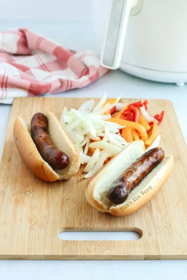 Brats in Air Fryer - side view of bratwurst in buns with onions and peppers behind on cutting board