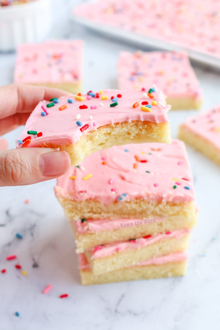 sugar cookie bars stacked up on top of each other. A hand is holding a single cookie bar with a bite removed above the stack.