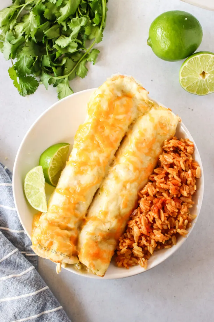 zoomed out image of two enchiladas on a plate with rice and sliced limes. Photo is from the top looking down.