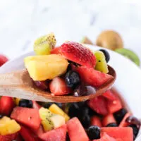 side view of fruit salad in bowl with a wooden spoon holding some above the bowl.