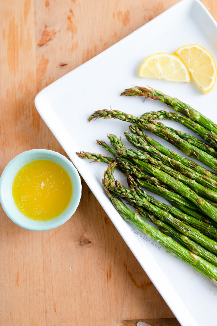 Cooked asparagus on a plate with a lemon wedge and small bowl of garlic butter..