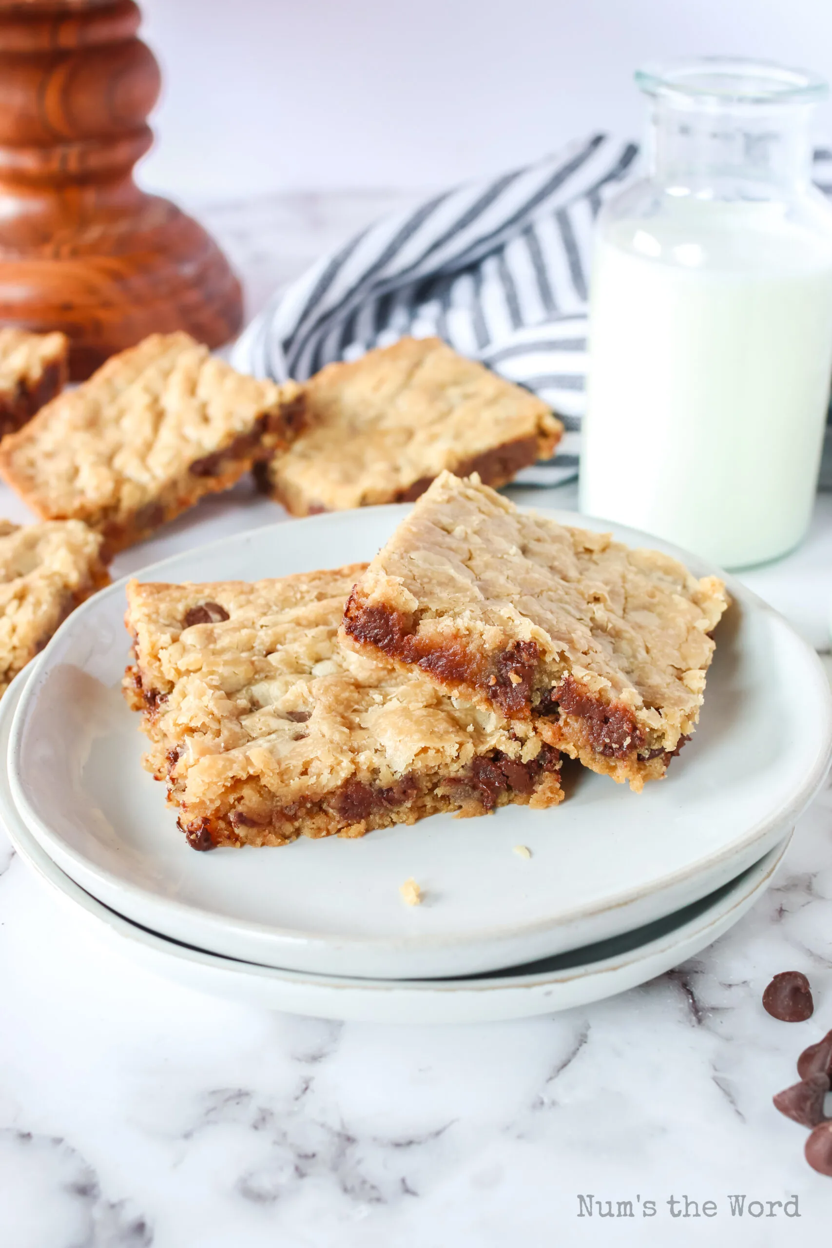 two cookie bars on a plate with a glass of milk and more cookie bars in the background.