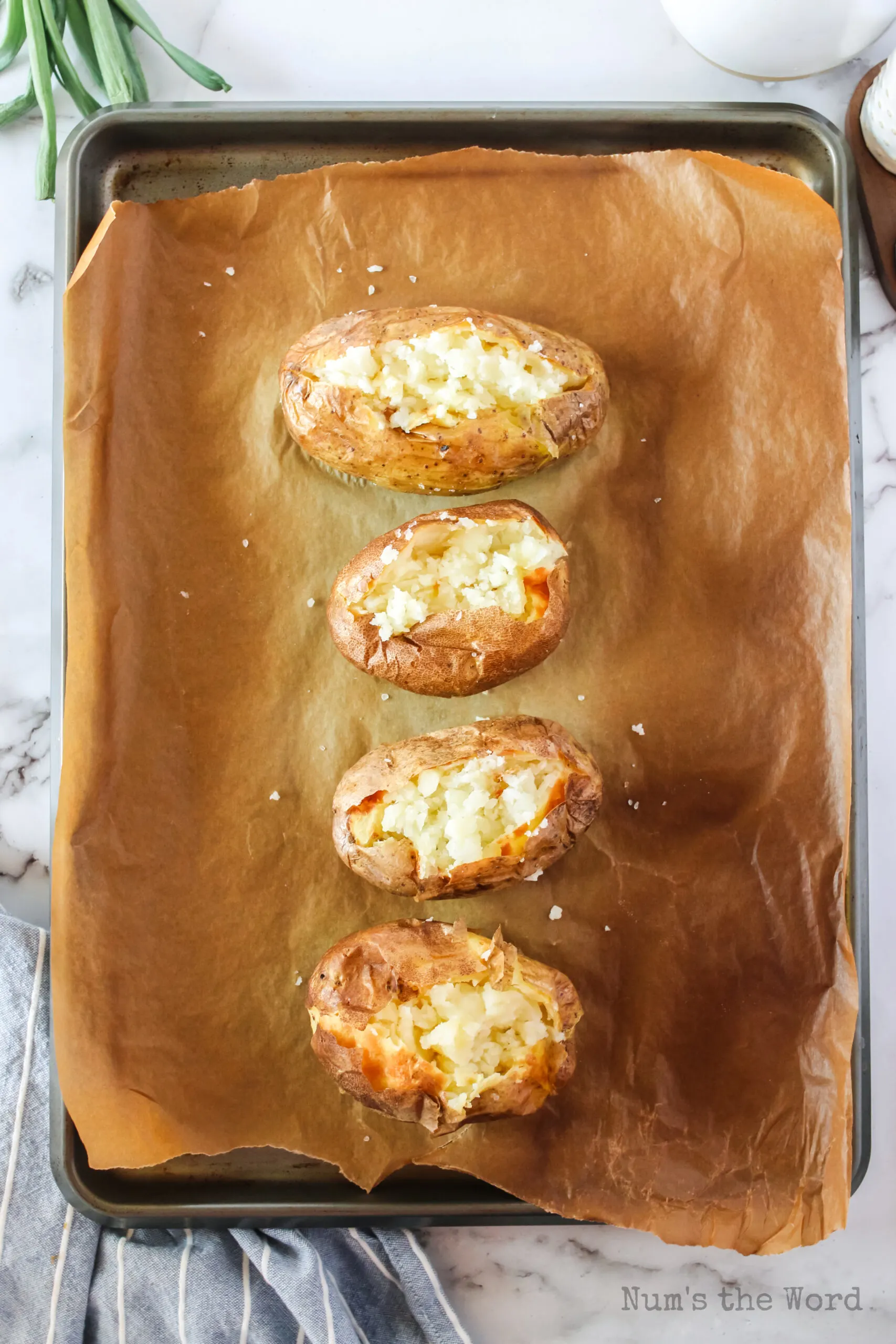 top view looking down, zoomed out image of baked potatoes on baking sheet. Potatoes are cut open to show off fluffy filling.