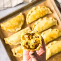 Baked empanadas on baking tray with a hand holding one cut open.