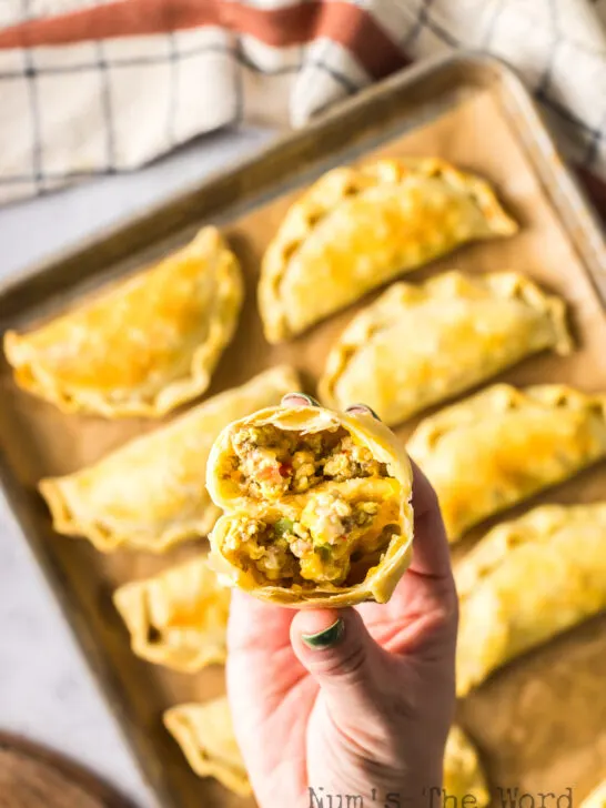 Baked empanadas on baking tray with a hand holding one cut open.