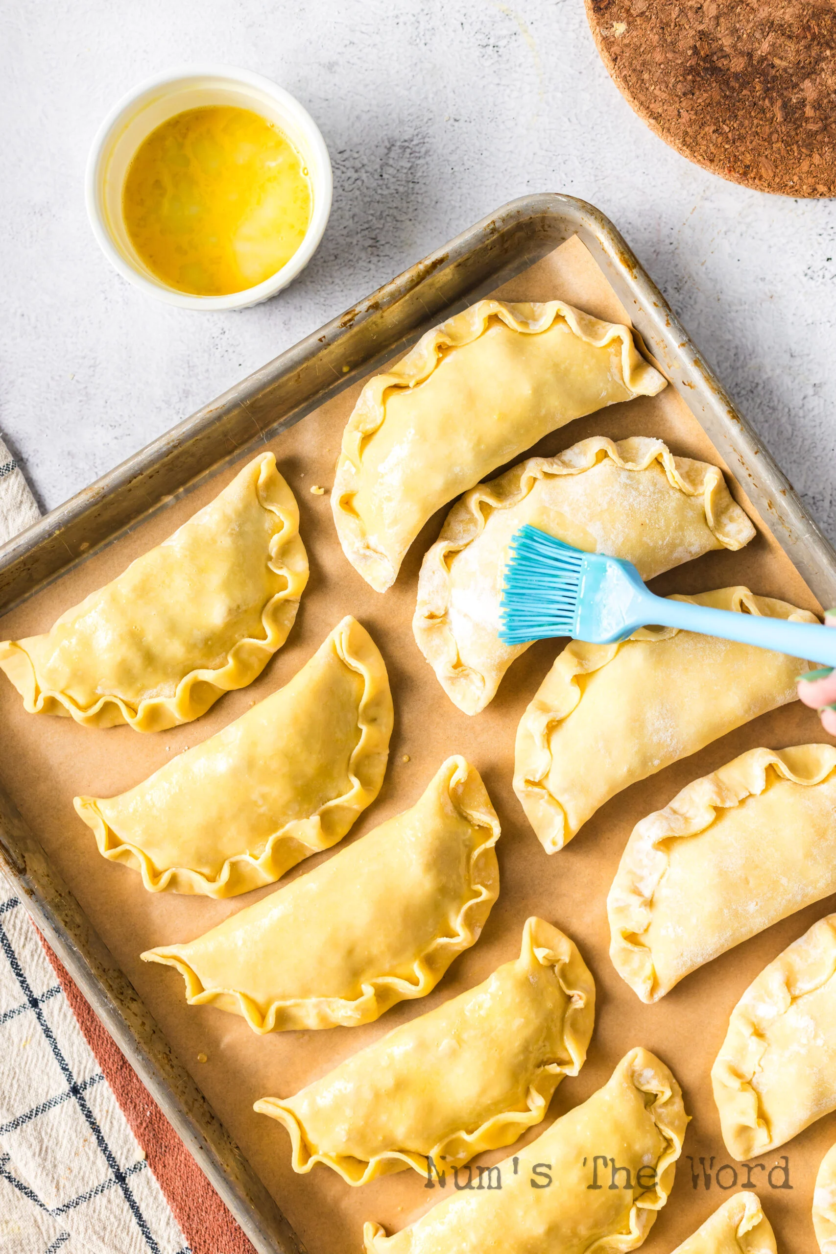 12 empanadas on a baking sheet being brushed with egg.